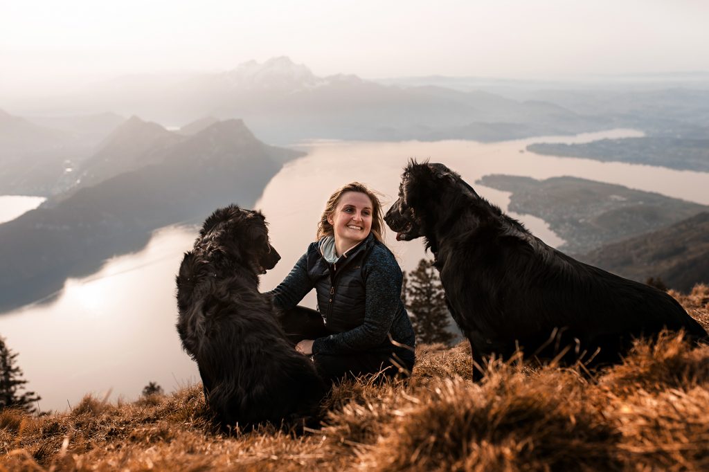 Zwei Flatcoated Retriever Hunde und eine junge Frau sitzen für ein Hundeshooting auf einem Berggipfel. Im Hintergrund ist der Vierwaldstättersee, der Pilatus, der Bürgenstock und das Stanserhorn zu sehen.