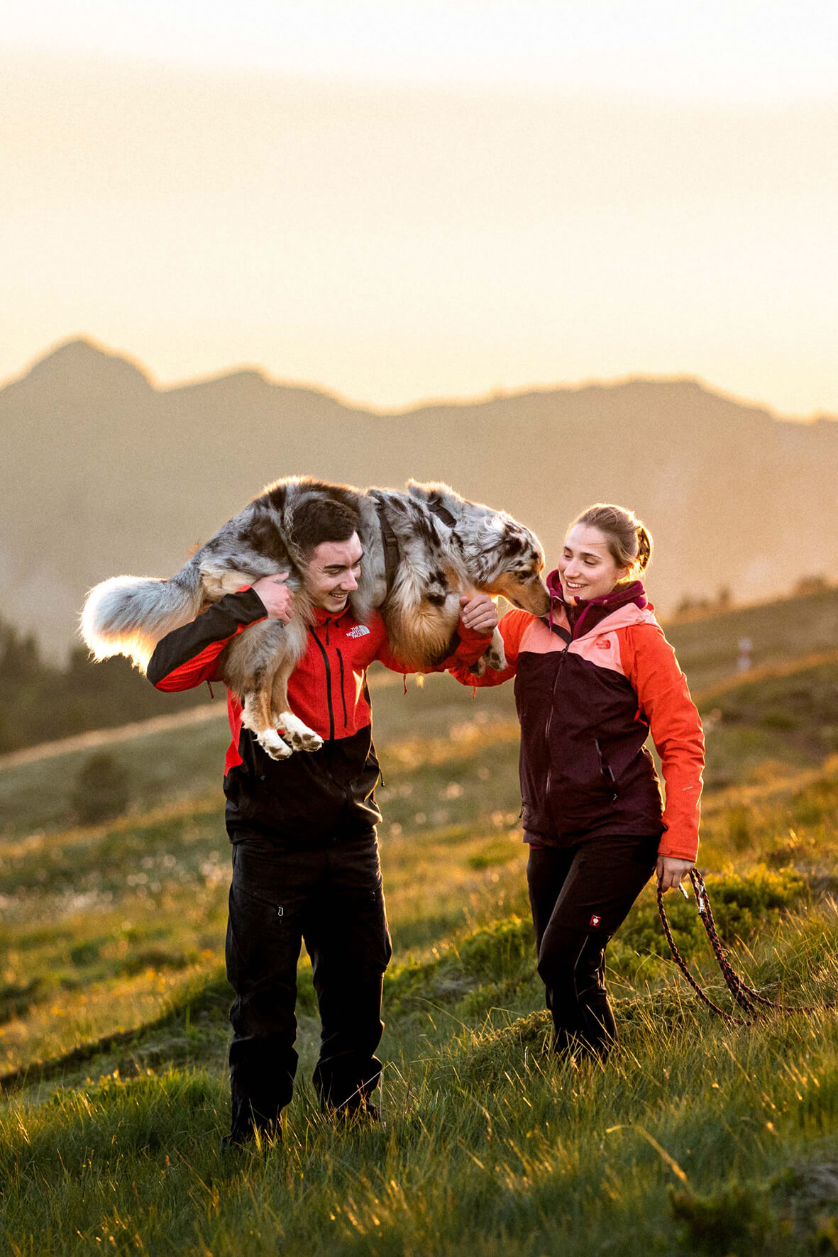 Ein Mann traegt einen Australian Shepherd auf seinen Schultern. Eine Frau unterstützt den Hund bei der Balance. Im Hintergrund ein Berg im rote Abendlicht. 