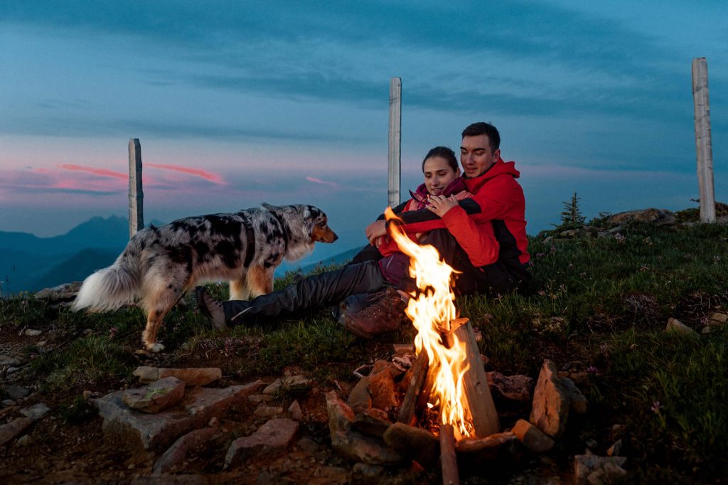 Ein Australian Shepherd und ein Paar sitzen an einem Lagerfeuer in den Bergen mit Sicht auf den Pilatus