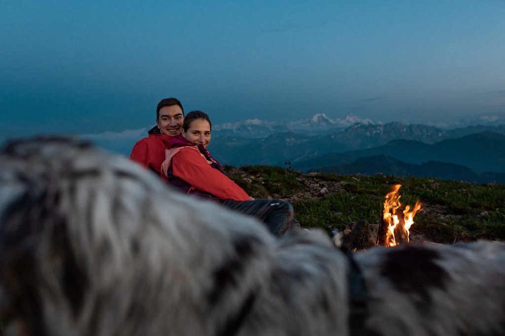 Ein Paar sitzt an einem Lagerfeuer in den Bergen. Im Hintergrund die Berner Alpen und im Vordergrund ein blue merle Australian Shepherd