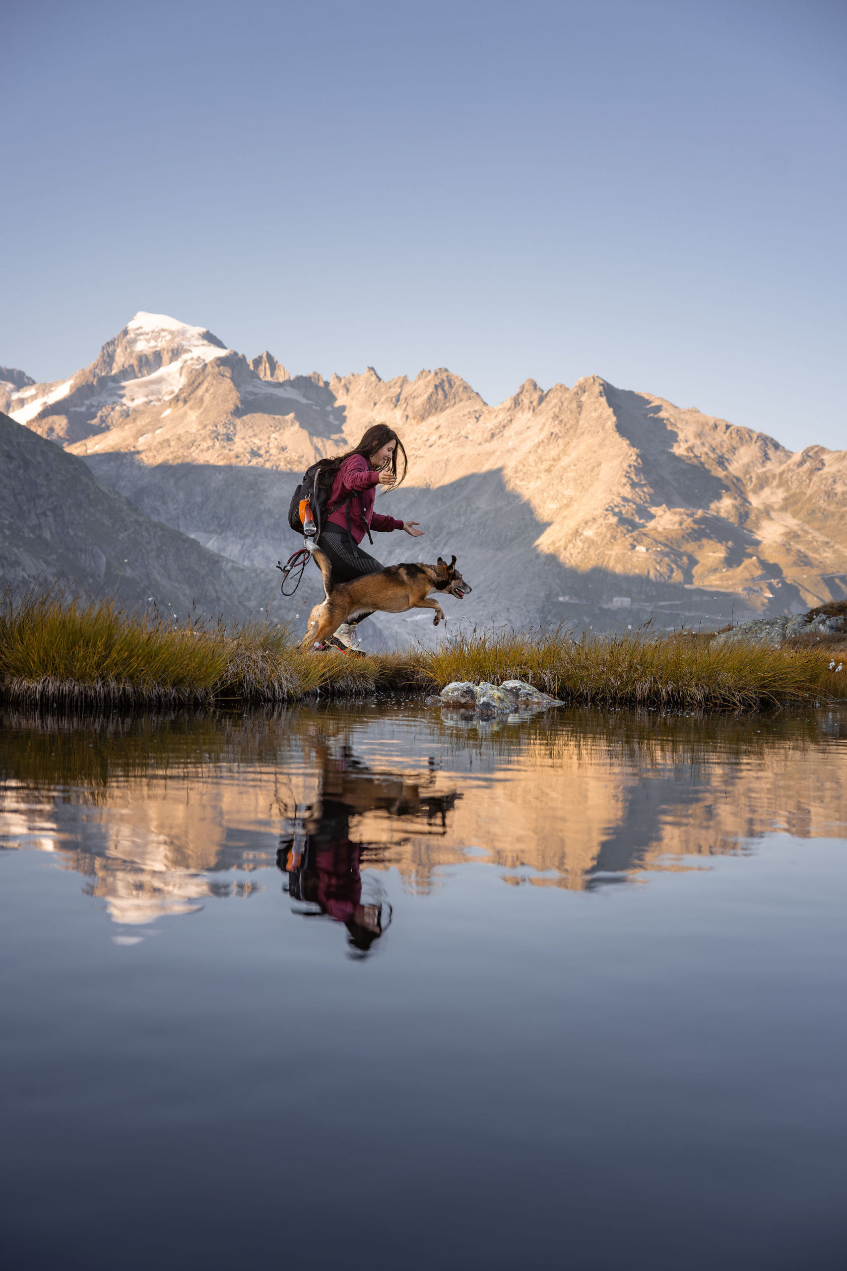Eine junge Frau und ein Hund springen über einen kleinen Bergsee, in dem sich der schneebedeckte Gipfel des berges im Hintergrund spiegelt. ebenfalls im Hintergrund ist der Furkapass der Schweiz zu sehen.